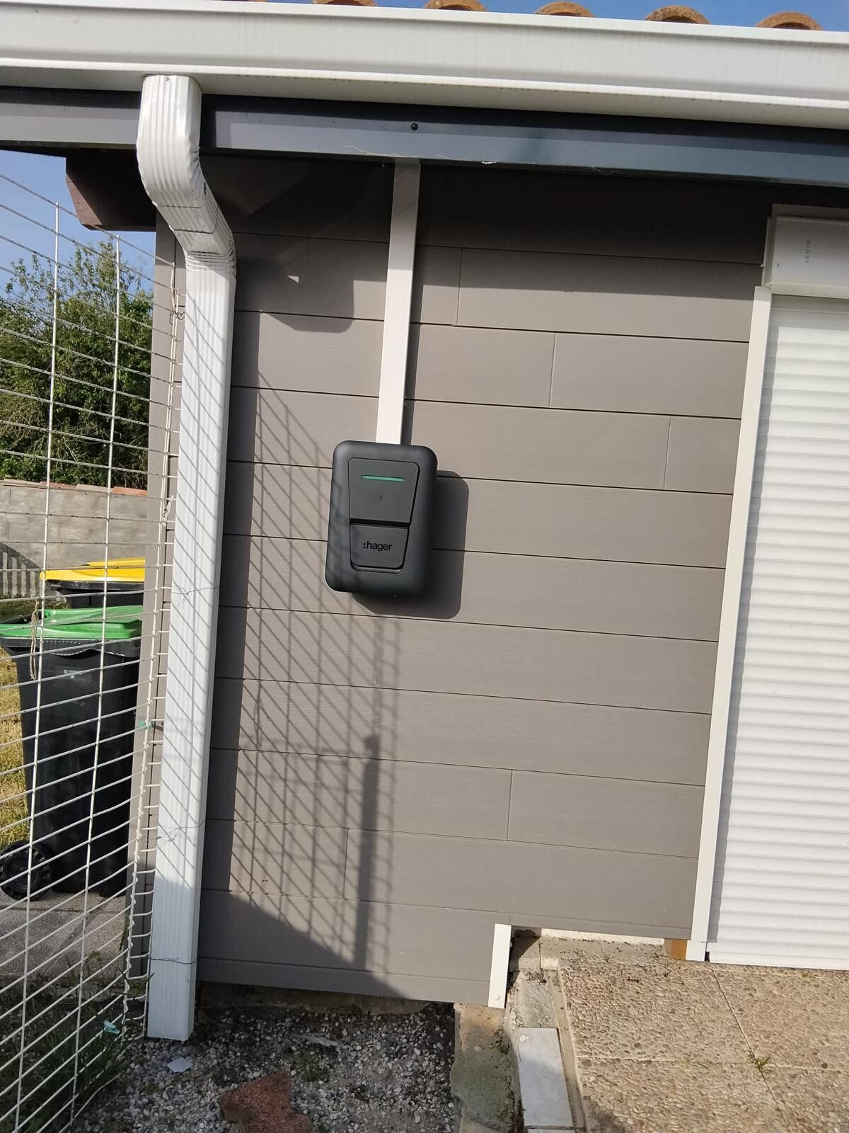 Borne de recharge murale posée sur un bardage gris, sous l'auvent d'un garage
