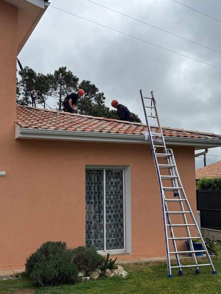 Deux poseurs casqués sur le toit d'une maison crépie orange, échelle contre la gouttière