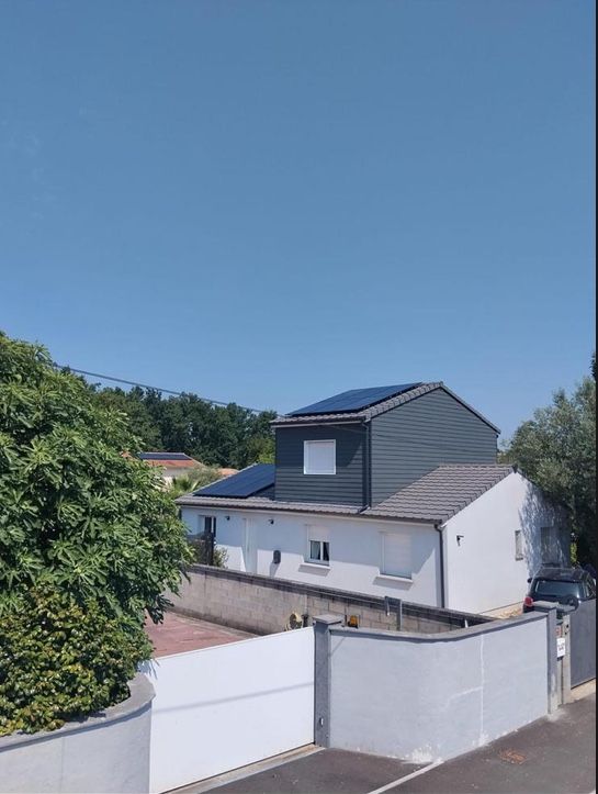 Maison blanche à étage en bardage gris, modules photovoltaïques noirs sur les deux pans de toit, ciel bleu