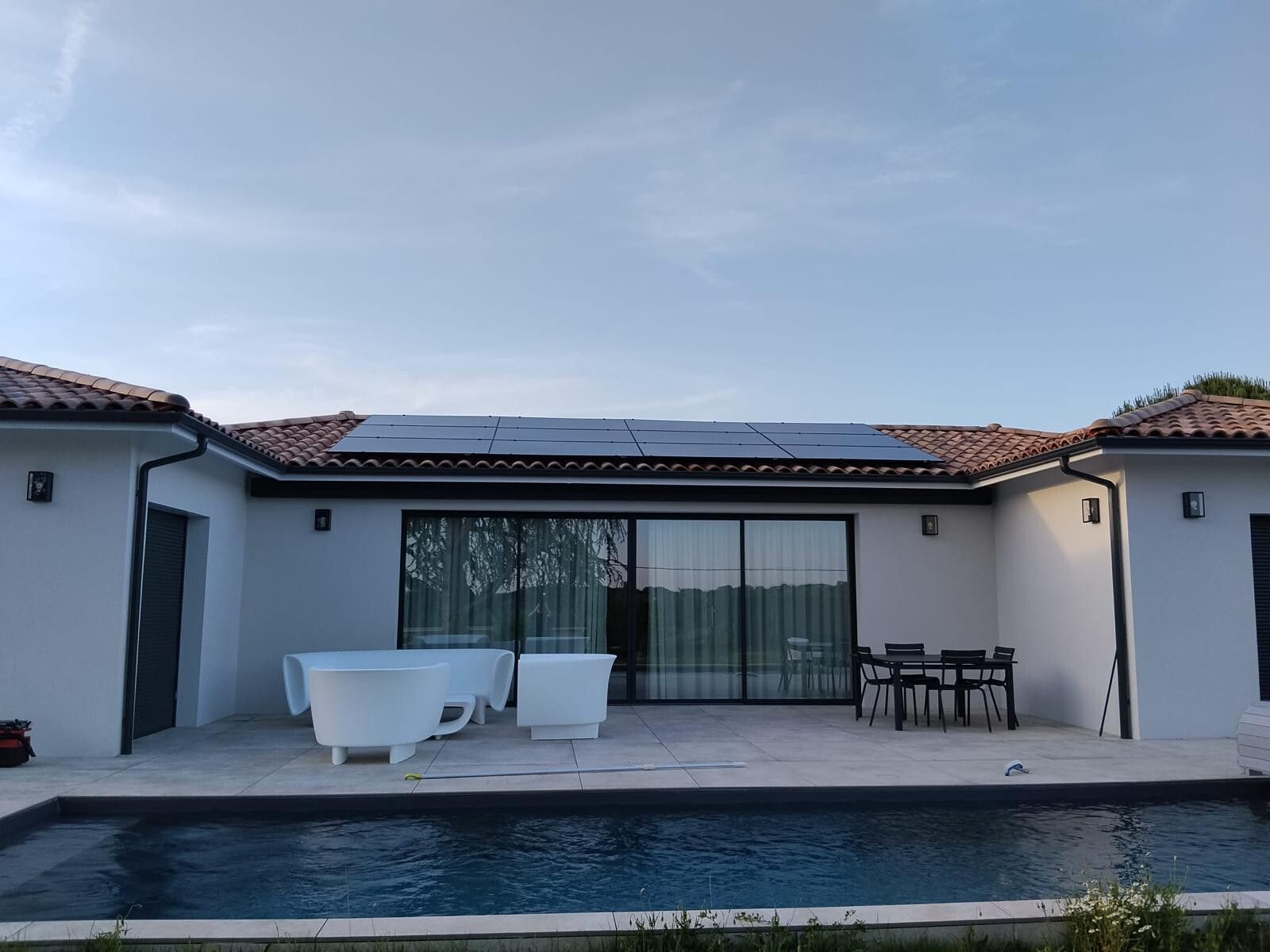 Maison de plain-pied avec ses modules photovoltaïques sur le toit, vue depuis la terrasse et la piscine en fin de journée